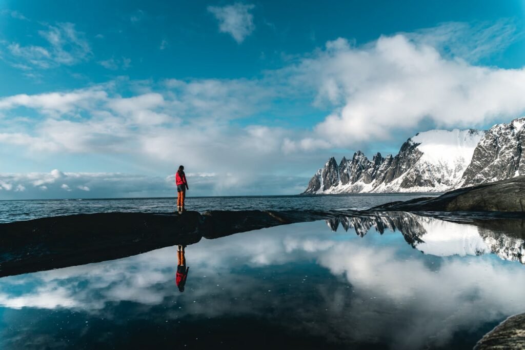 A person stands by a scenic snowy coastline reflected in still waters, under a vibrant blue sky.