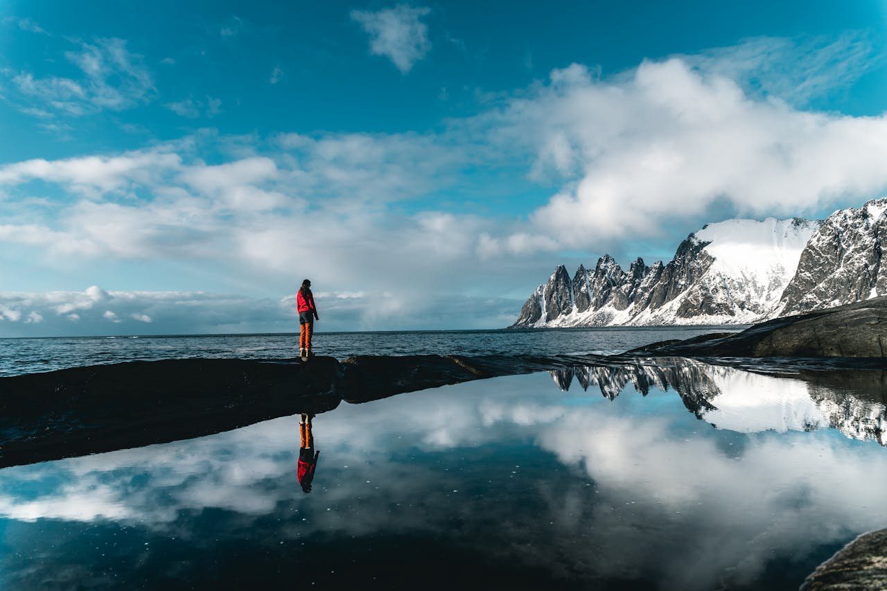 A person stands by a scenic snowy coastline reflected in still waters, under a vibrant blue sky.